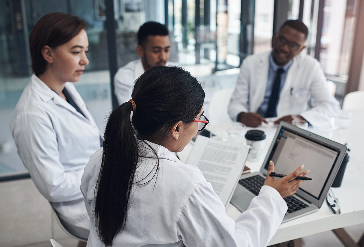 Four researchers in a meeting around a table, reviewing documents and discussing information displayed on a laptop in an office.